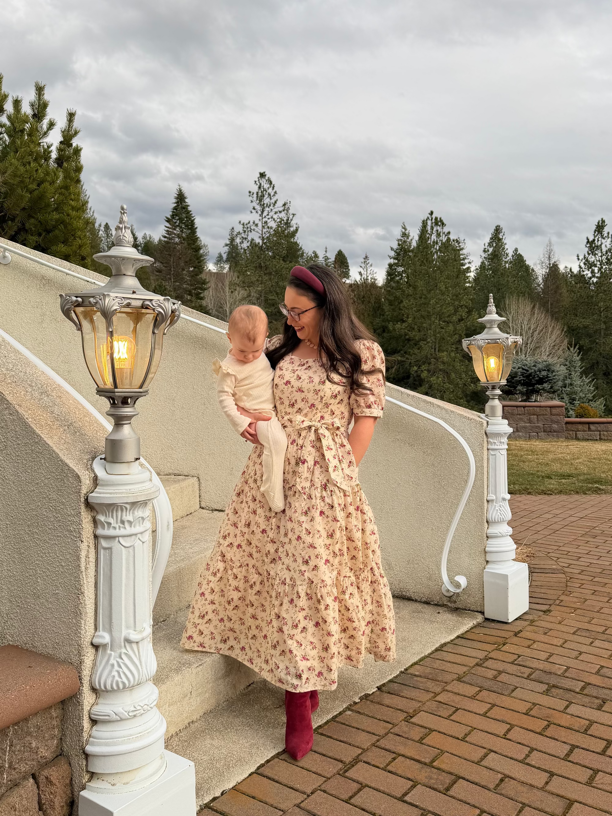 Woman in a floral dress holding a baby on a staircase with a cloudy sky and trees in the background.