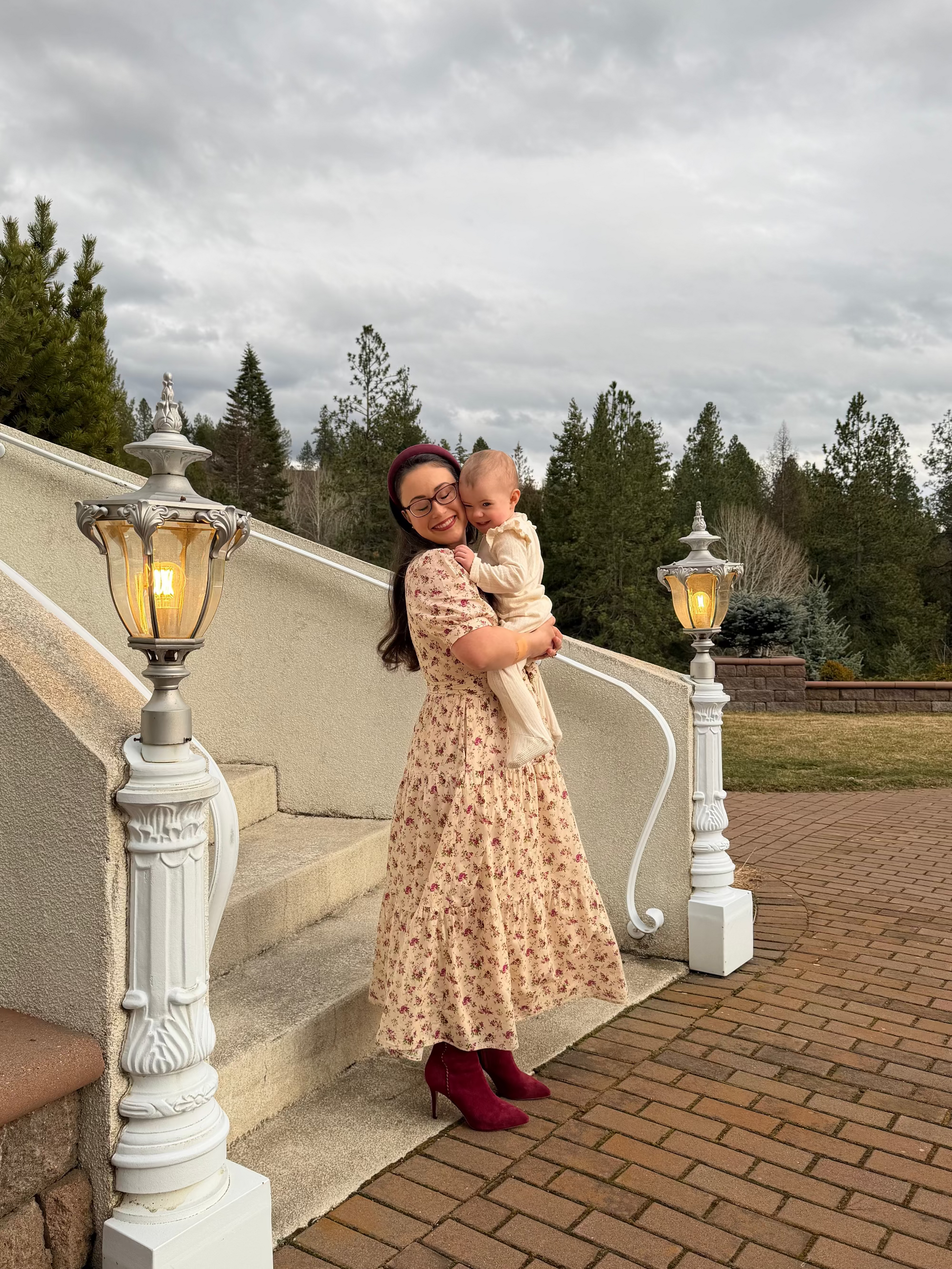 Woman in a floral dress holding a baby on a staircase with decorative lamps and trees in the background.