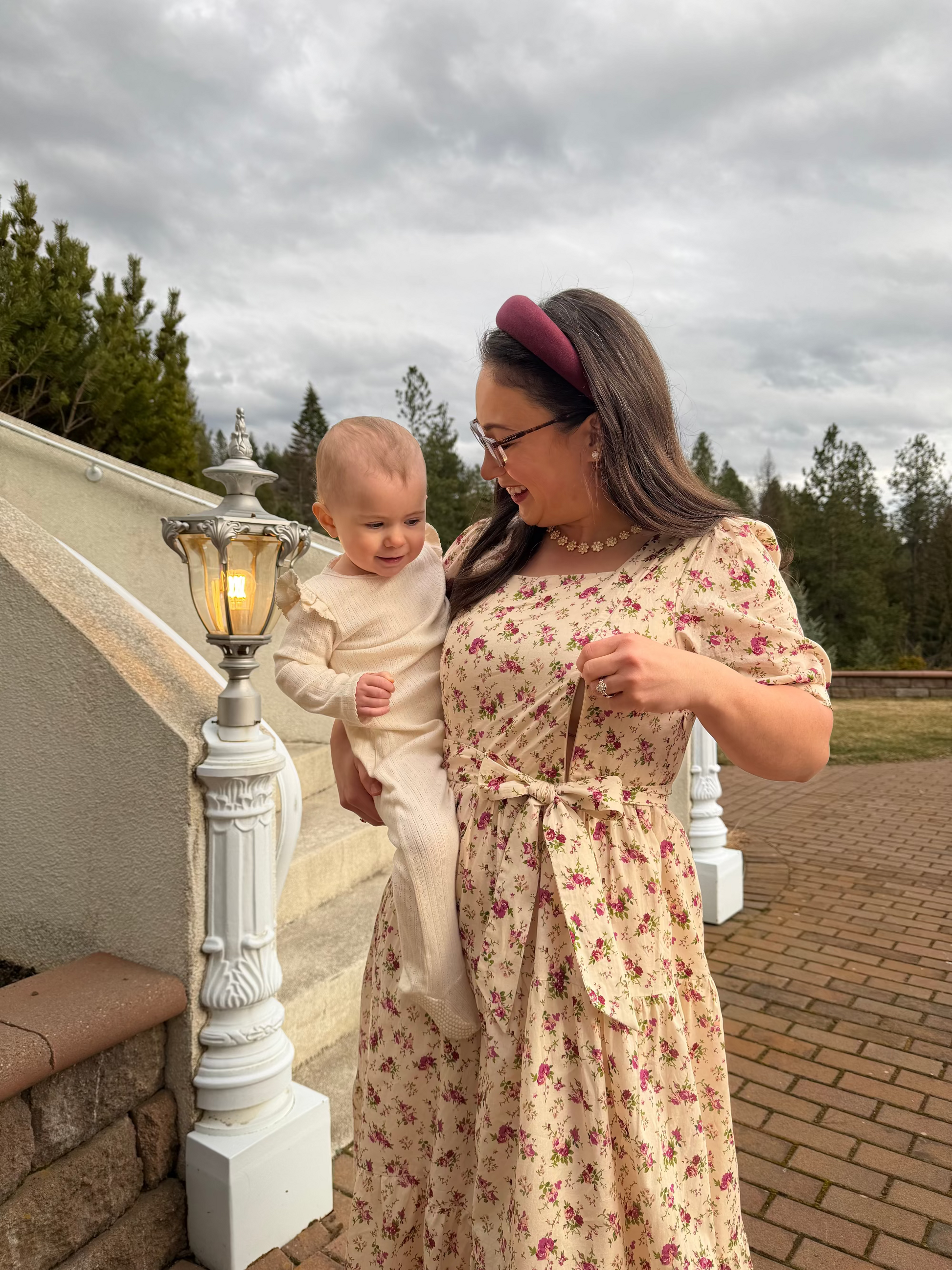 Woman in a floral dress holding a baby outdoors with a cloudy sky.